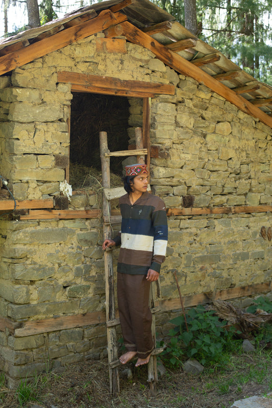 Model wearing a panelled linen embroidered shirt standing on a wooden ladder in front of a stone building with a wooden roof.