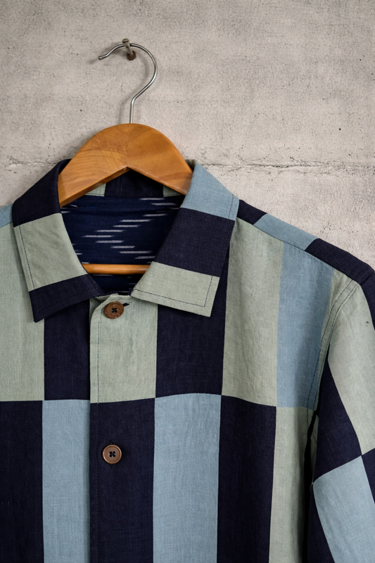 Close up of patchwork checkered linen shirt with dark blue, light green, and light blue pattern displayed against a wall