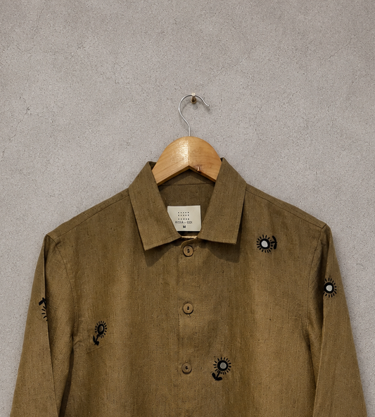A brown linen button-up shirt with mirror embroidered details, hanging on a wooden hanger against a textured gray wall.