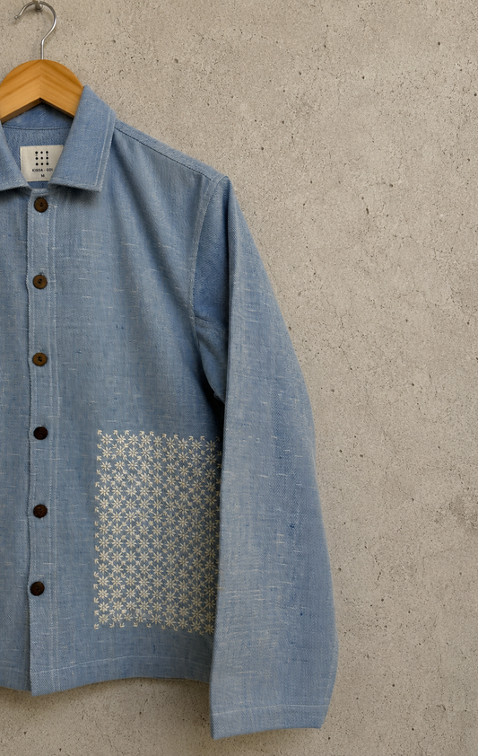 A light blue textured linen jacket with dark buttons and a geometric patterned embroidery,hanging on a wooden hanger against a textured concrete wall.