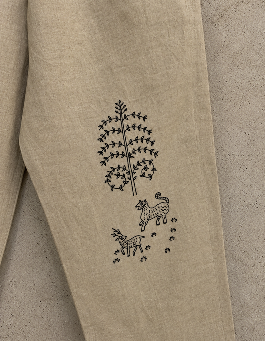 Close-up of a beige linen garment laid flat on a textured beige surface, showing a black embroidered tree and deer motif on the fabric.