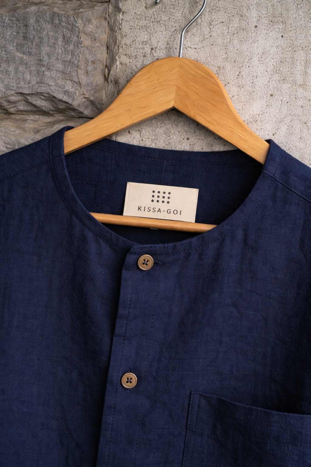 Closeup of blue collarless linen shirt displayed against a grey wall