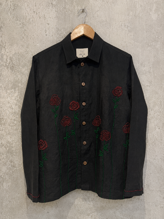 A black long-sleeve button-up shirt with embroidered red roses and green stems on the front, hanging on a wooden hanger against a textured gray wall.