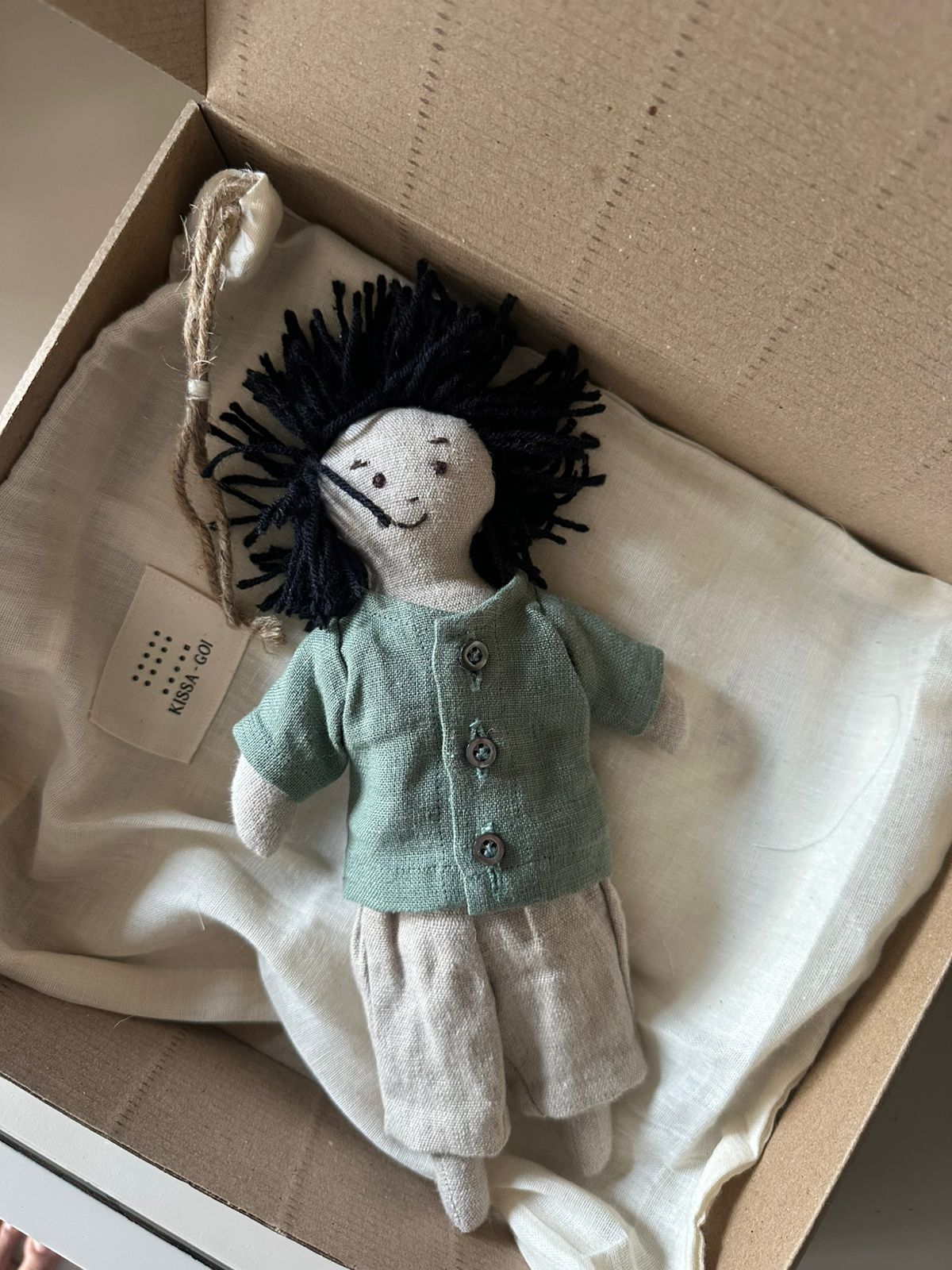 Stuffed doll with a solid linen shirt and white linen pants on a white bag inside a cardboard box. The first friend hand-stitched doll