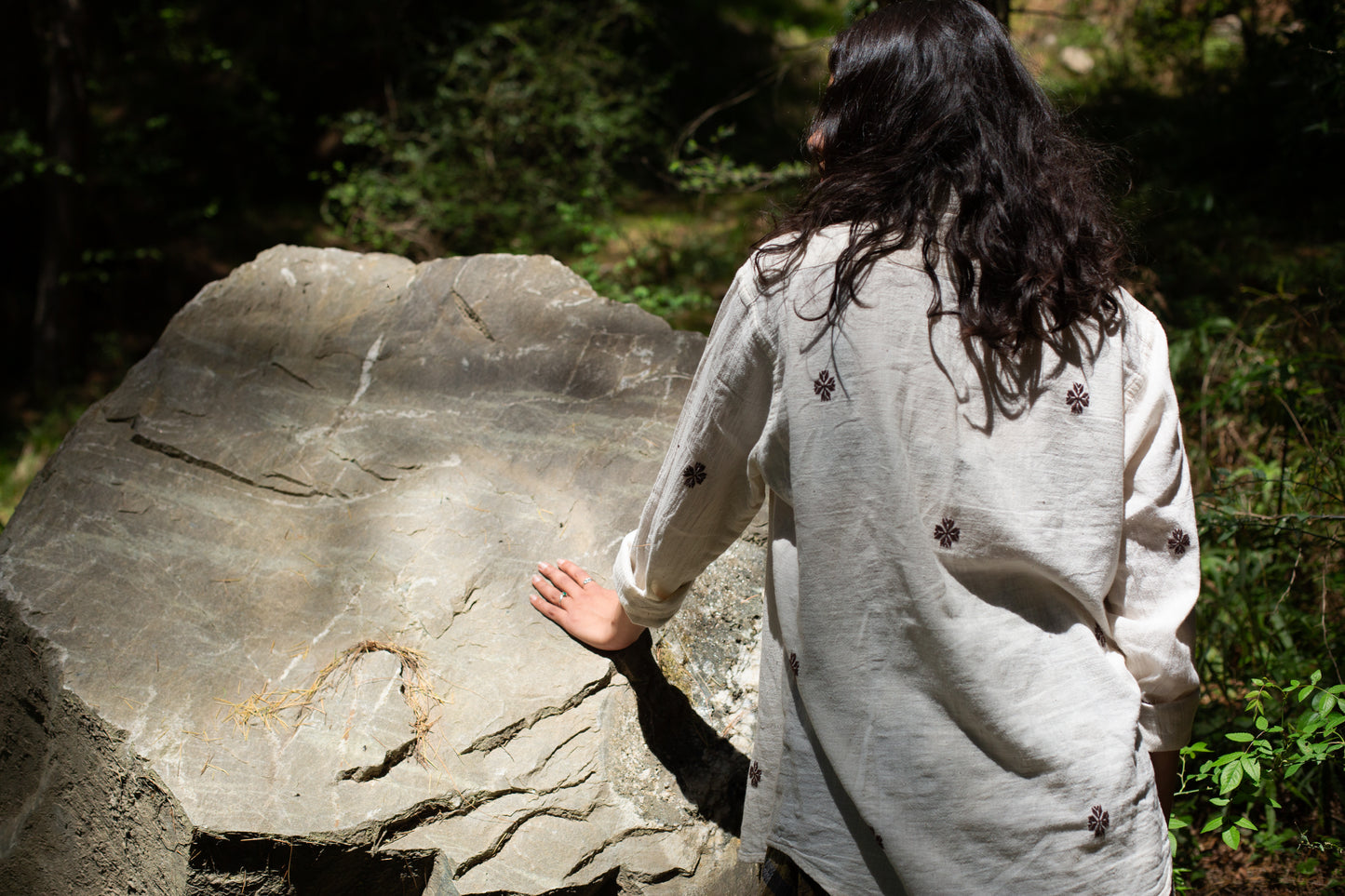 Person in a white cotton shirt with traditional suf embroidery standing next to a large rock in a forest
