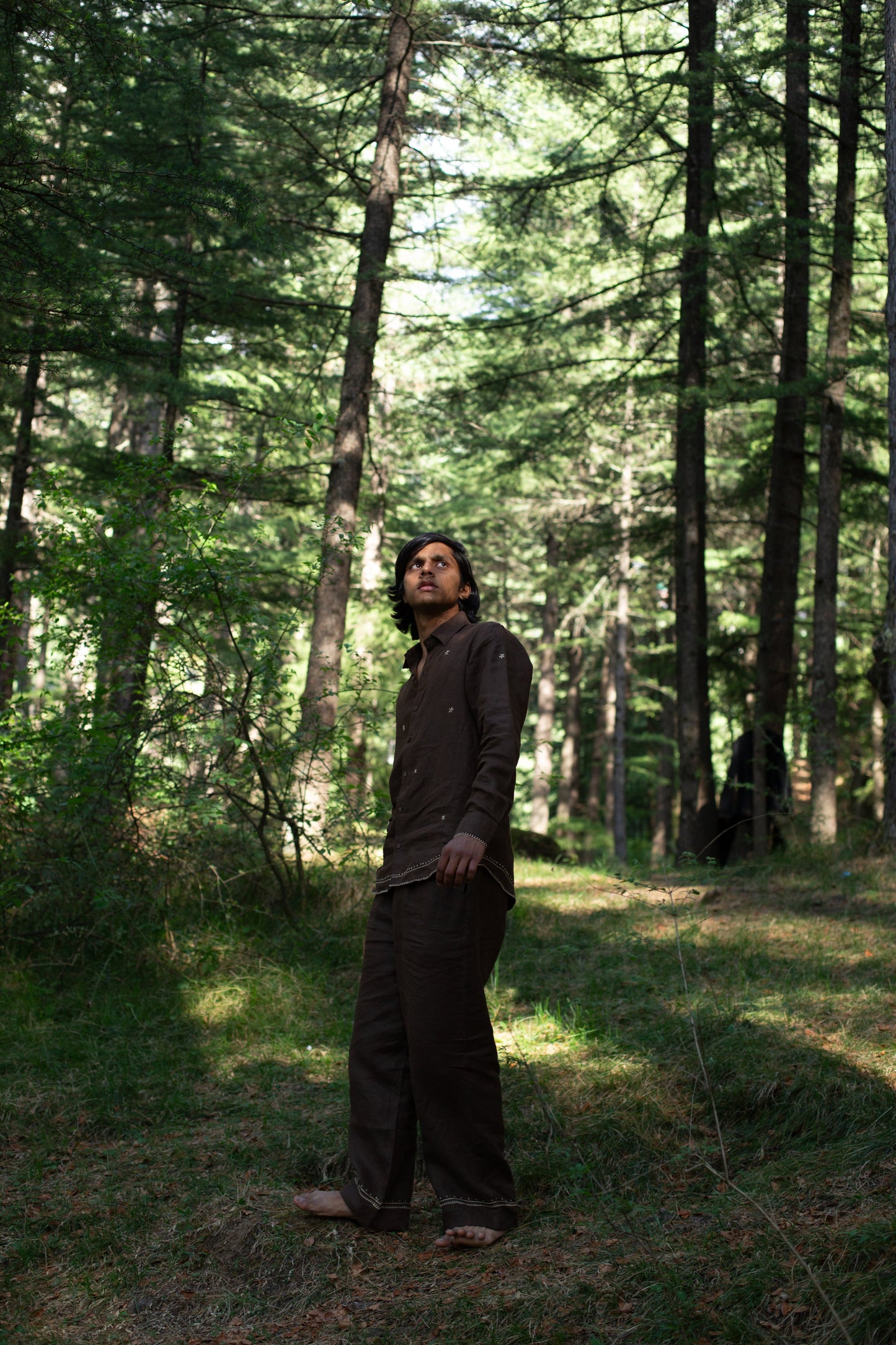 Model wearing a brown embroidered linen shirt standing in a forest with tall trees and dappled sunlight.