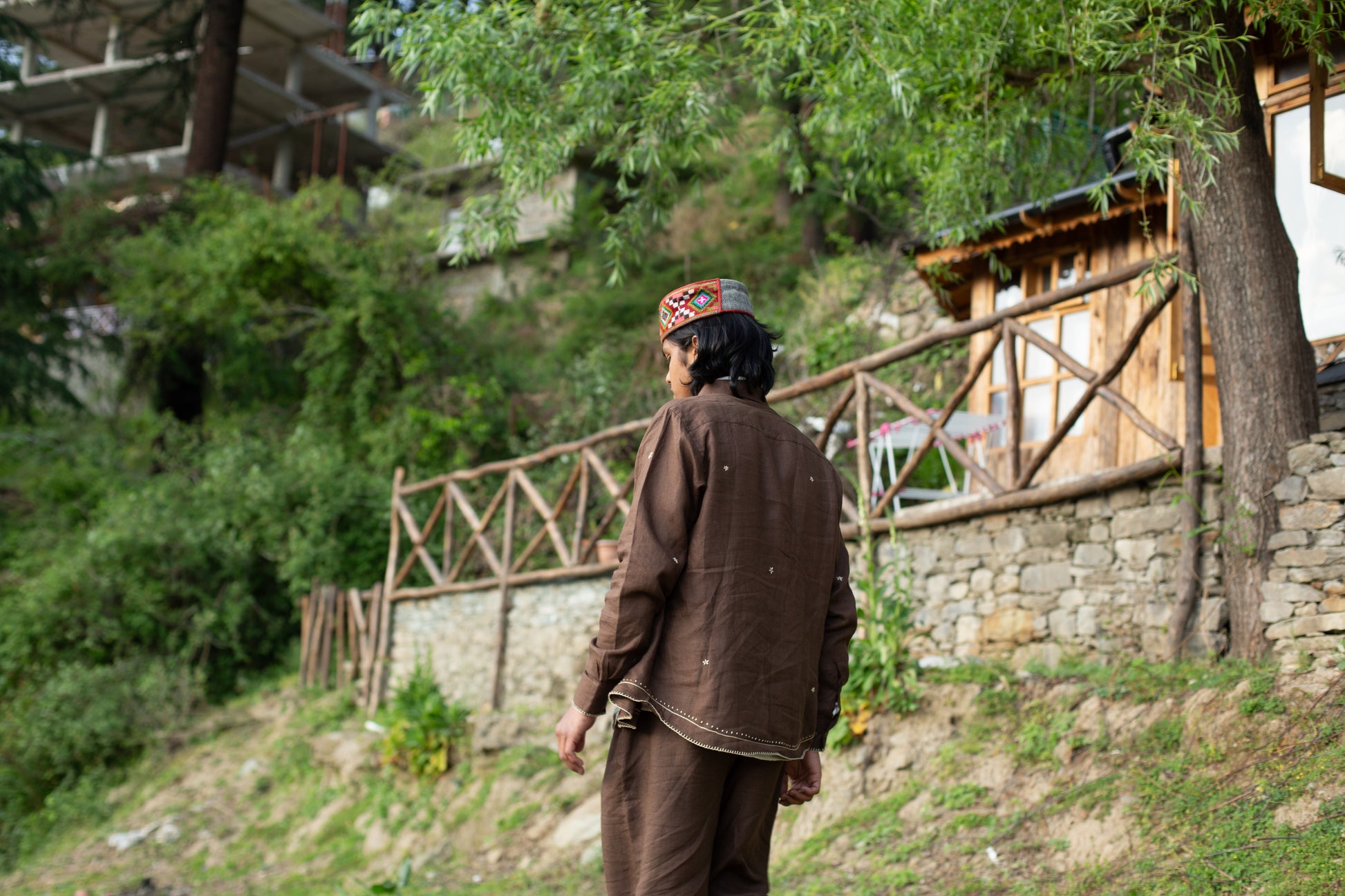 Model wearing brown embroidered linen shirt standing in a natural setting with wooden structures and greenery.