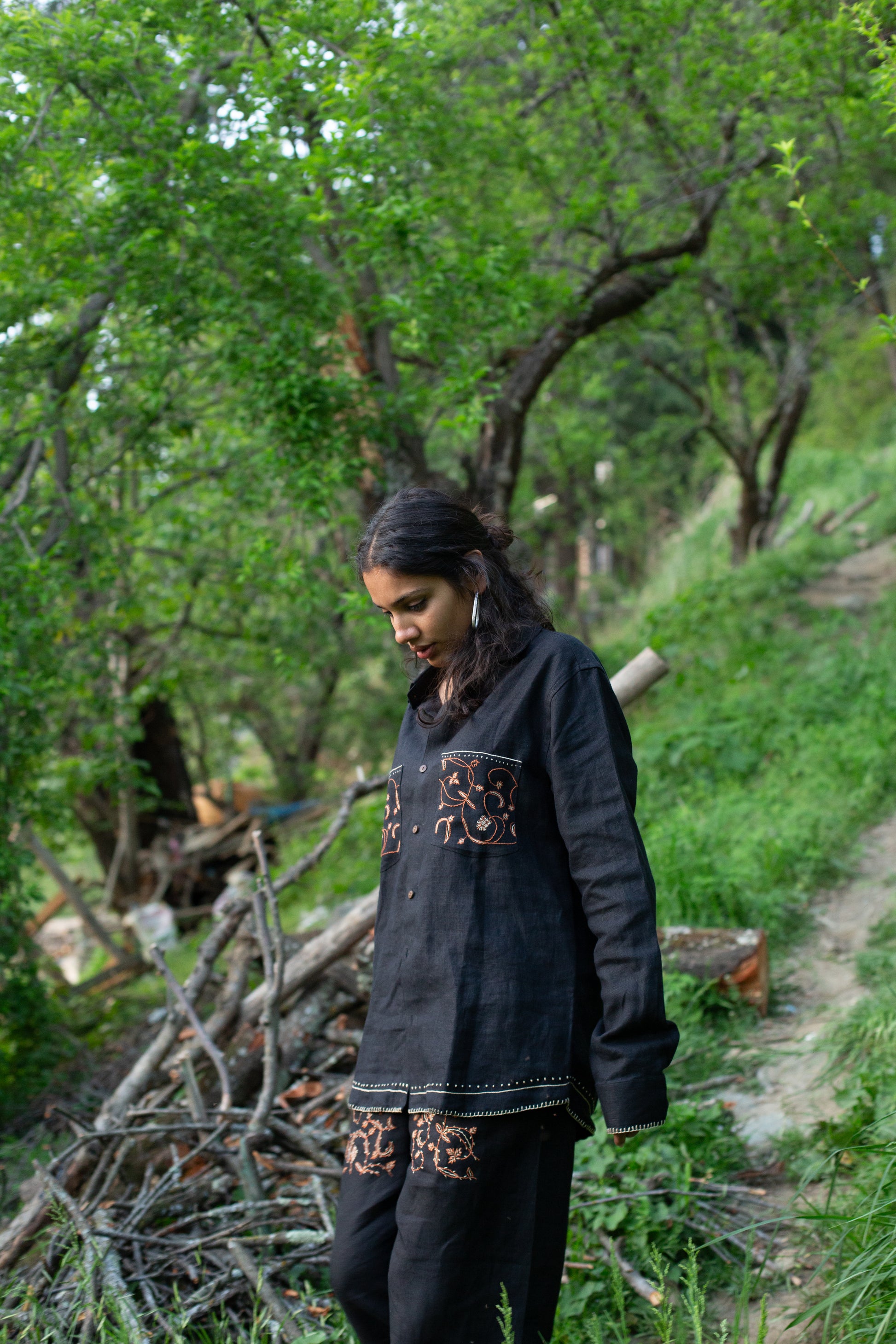 Model wearing a black linen shirt with embroidered pockets and a patterned design in a forest setting