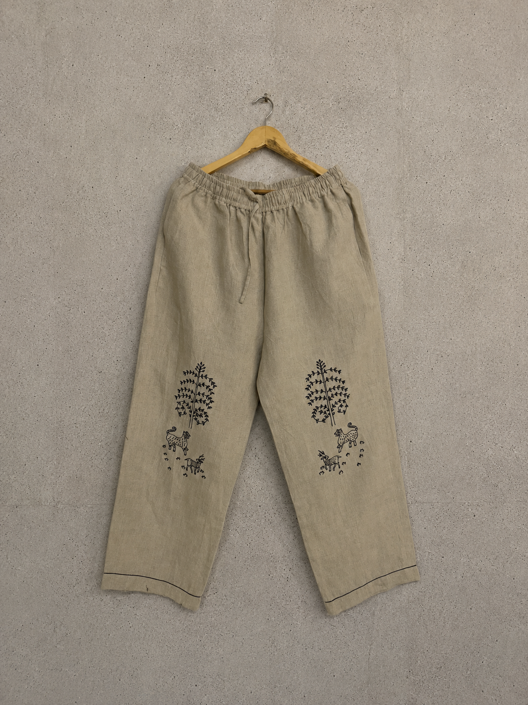Beige linen drawstring pants with black embroidered floral motifs on the lower legs, hanging on a wooden hanger against a textured gray wall.