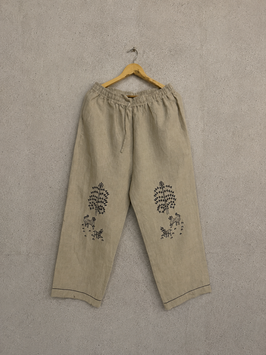 Beige linen drawstring pants with black embroidered floral motifs on the lower legs, hanging on a wooden hanger against a textured gray wall.