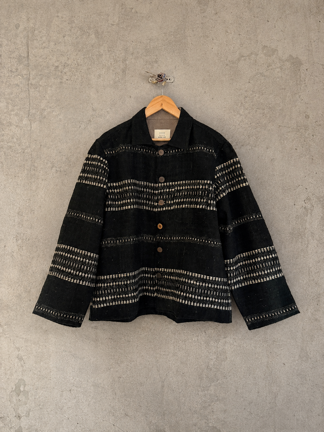 A black woven button-up woollen jacket with horizontal off-white textured stripe patterns, hanging on a wooden hanger against a textured gray concrete wall.