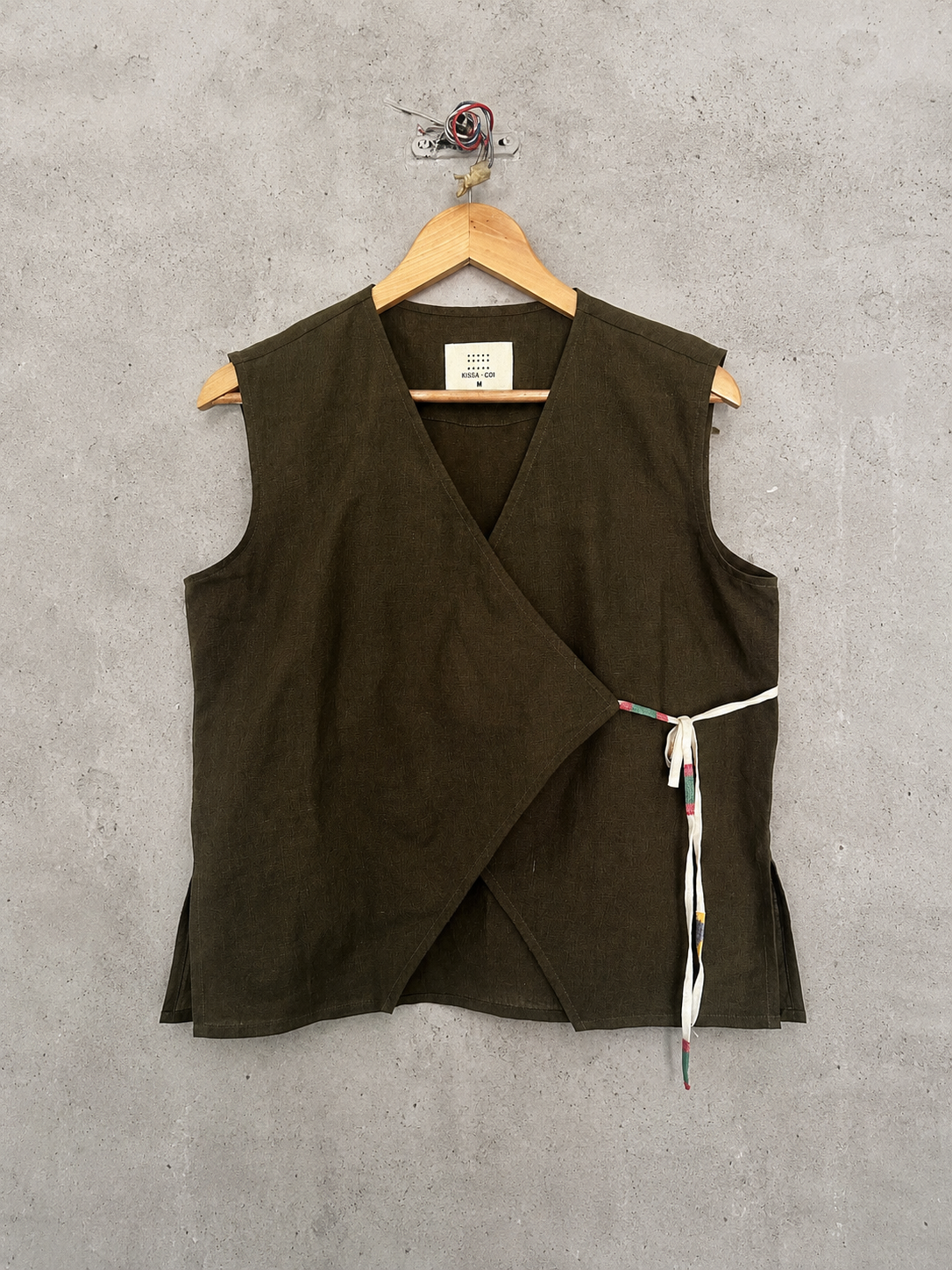 Olive vest wrap top with a tie detail on a hanger against a gray concrete wall.