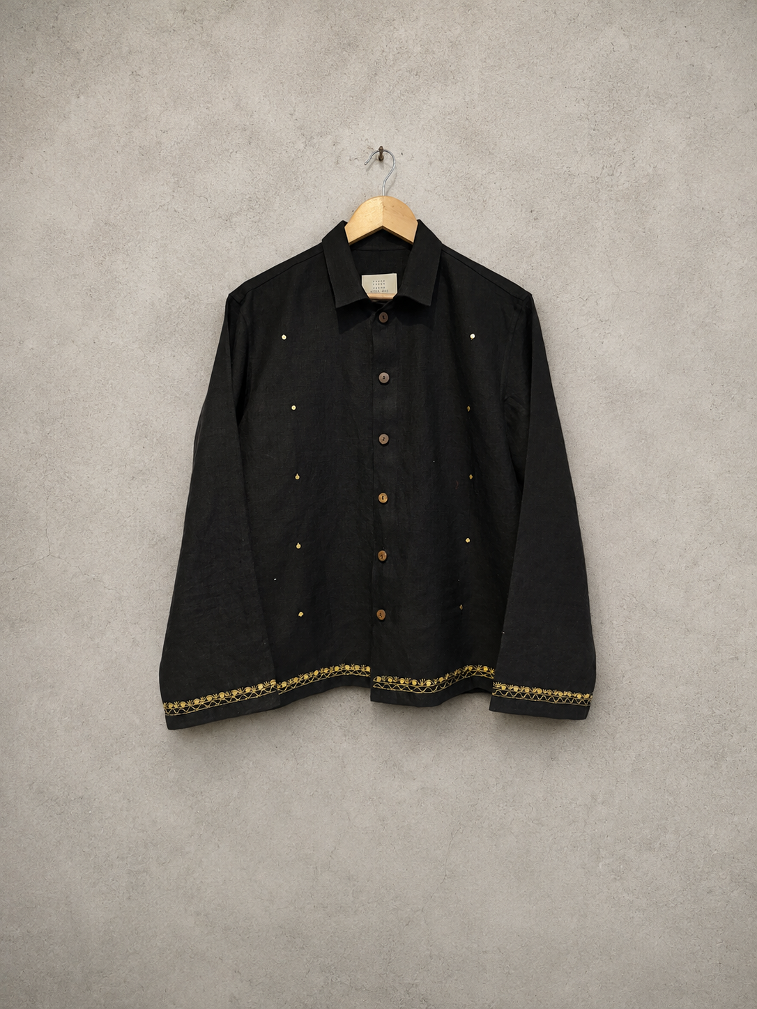 A black long-sleeve button-up shirt with gold embroidered details and gold buttons, hanging on a wooden hanger against a textured gray wall.