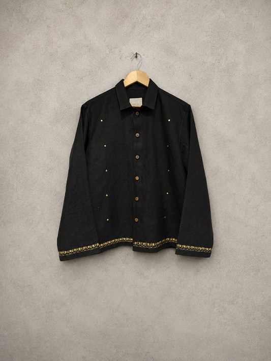 A black long-sleeve button-up shirt with gold embroidered details and gold buttons, hanging on a wooden hanger against a textured gray wall.