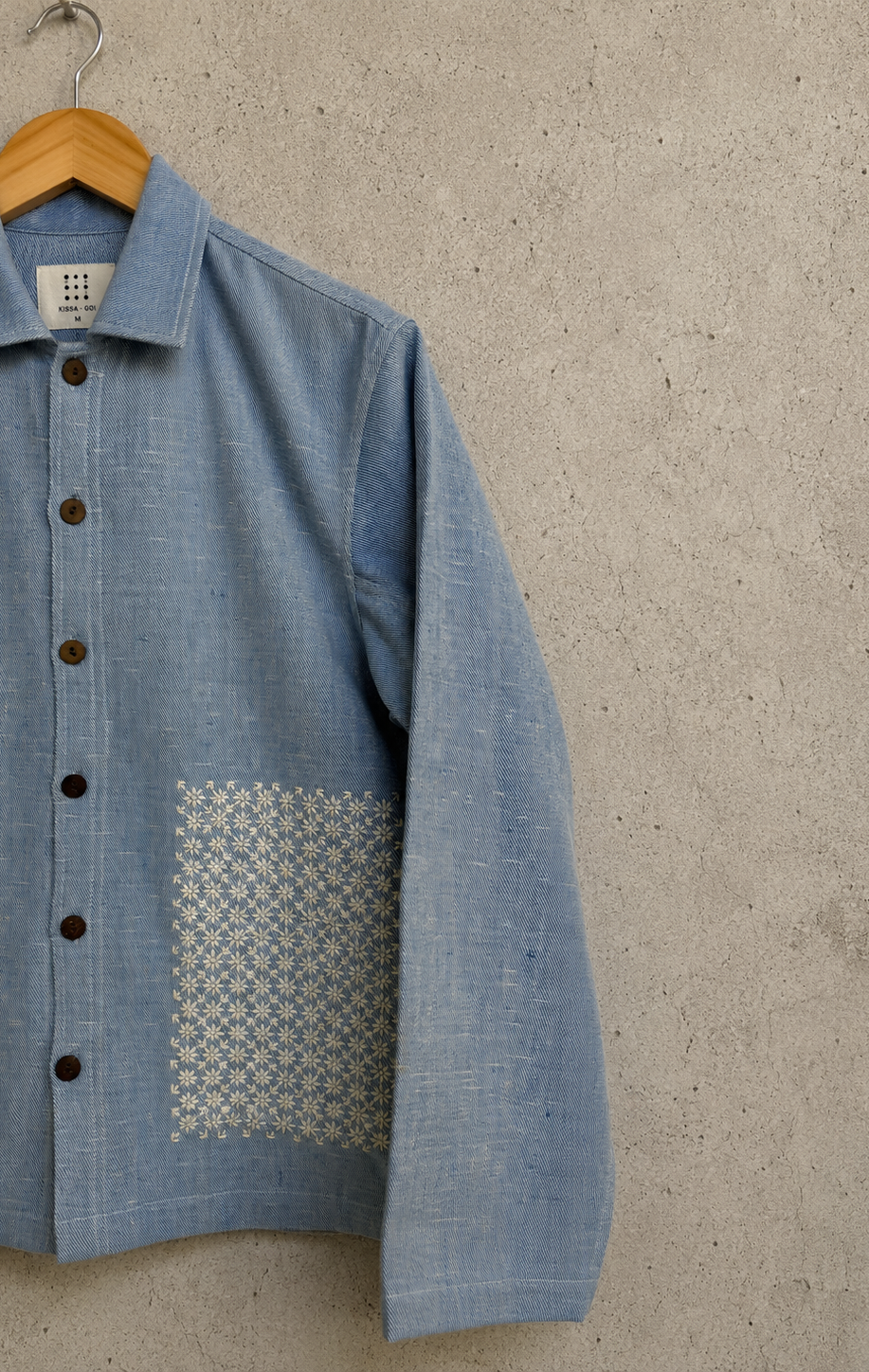 A light blue textured linen jacket with dark buttons and a geometric patterned embroidery,hanging on a wooden hanger against a textured concrete wall.