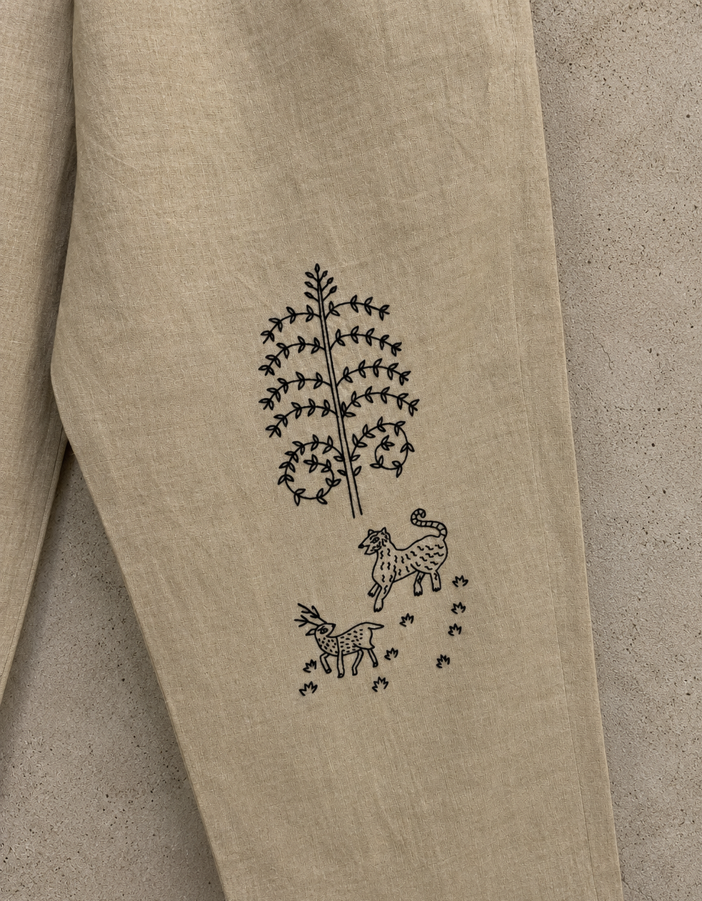 Close-up of a beige linen garment laid flat on a textured beige surface, showing a black embroidered tree and deer motif on the fabric.