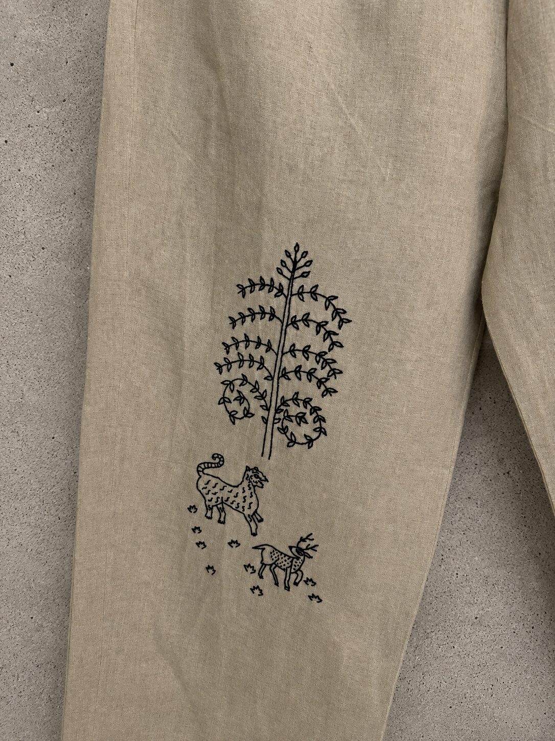Close-up of a beige linen garment laid flat on a gray concrete surface, featuring a black embroidered design of a tree, a tiger and deer on the fabric.