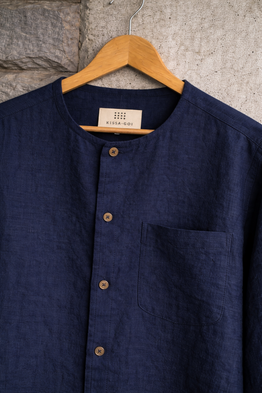 Closeup of blue collarless linen shirt displayed against a grey wall