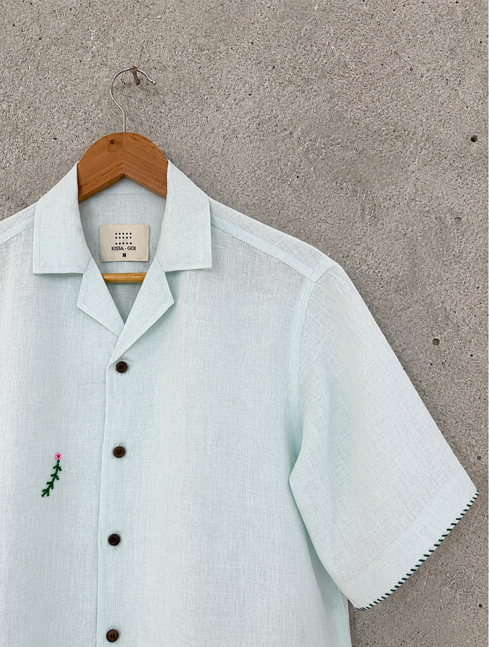A pale powder blue short-sleeve button-up shirt with a pointed collar and a small embroidered green plant with a pink flower on the chest, hanging on a wooden hanger against a textured gray wall.
