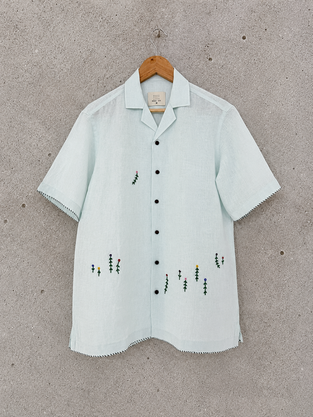 A light blue short-sleeve button-up shirt with delicate floral embroidery, hanging on a wooden hanger against a textured gray concrete wall.