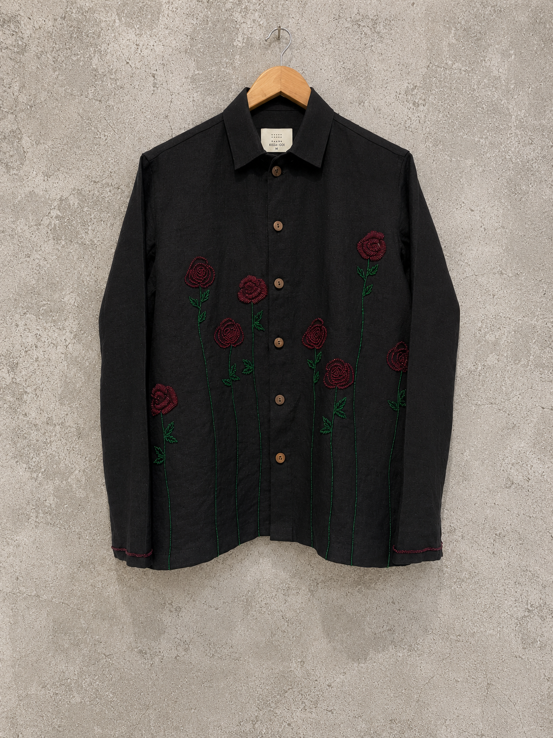 A black long-sleeve button-up shirt with embroidered red roses and green stems, hanging on a wooden hanger against a textured gray concrete wall.