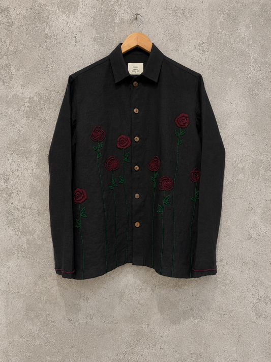 A black long-sleeve button-up shirt with embroidered red roses and green stems, hanging on a wooden hanger against a textured gray concrete wall.
