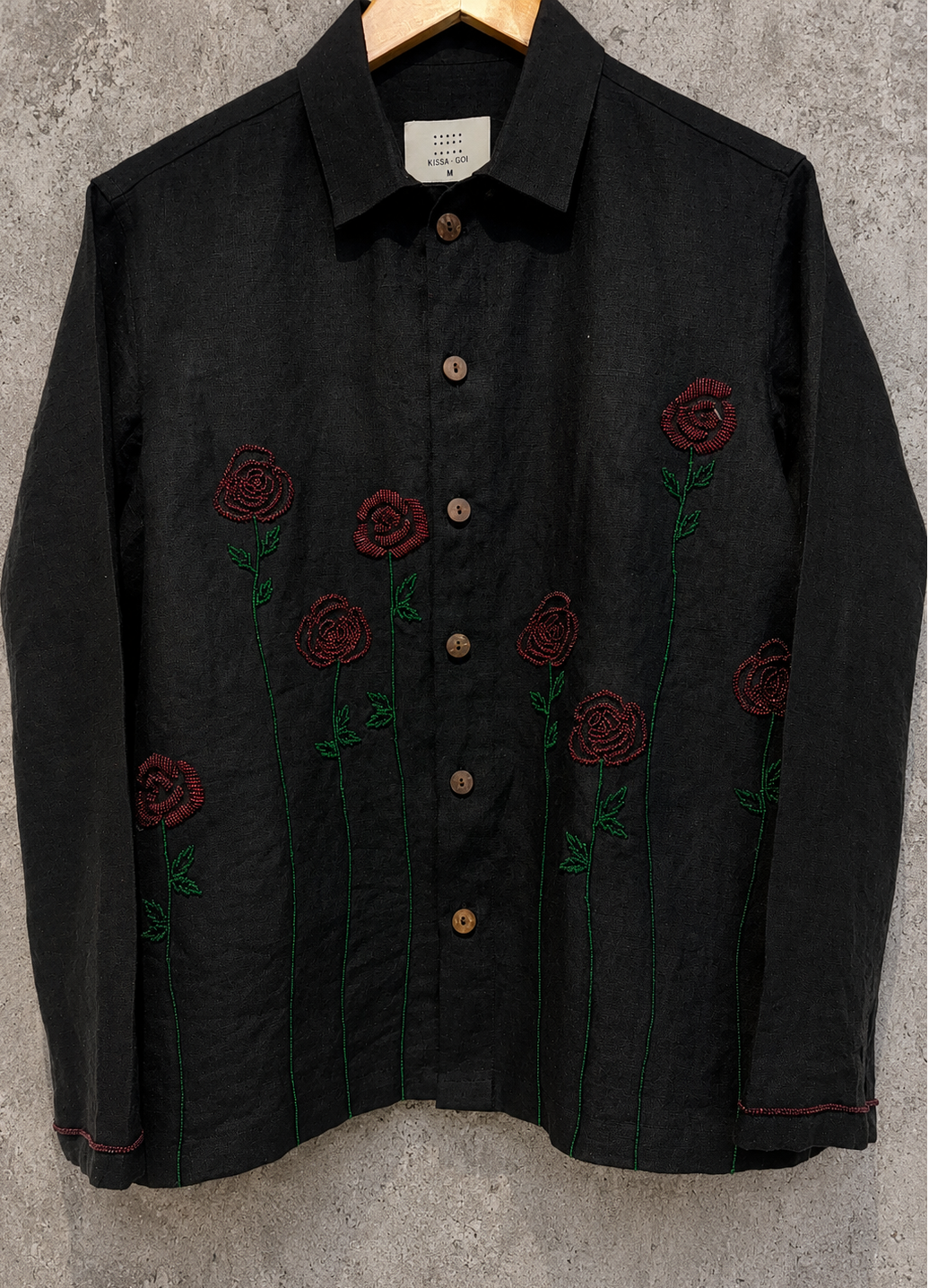 A black long-sleeve button-up shirt with embroidered red roses and green stems on the front, hanging on a wooden hanger against a textured gray wall.