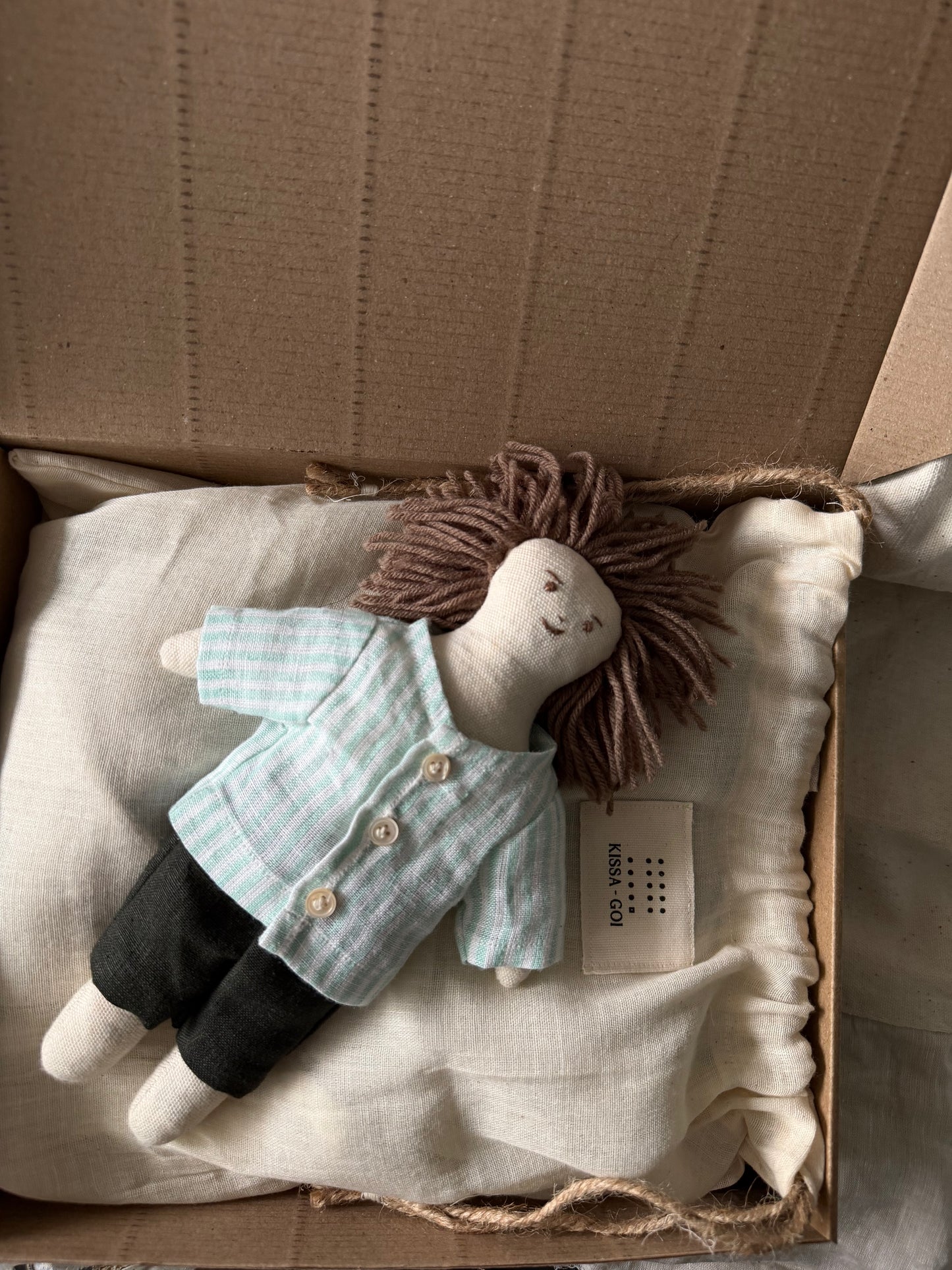 Stuffed doll with a striped shirt and dark pants on a white bag inside a cardboard box. The first friend hand-stitched doll