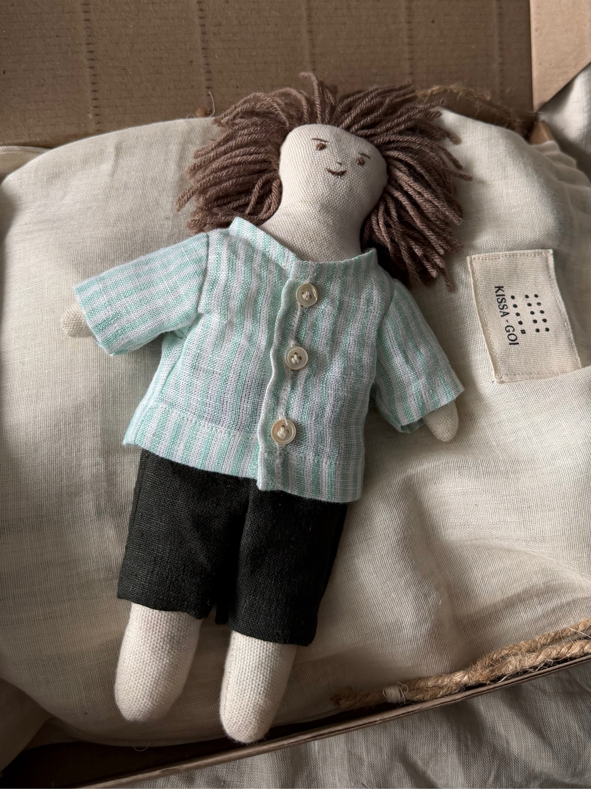 Stuffed doll with a striped shirt and dark pants on a white bag inside a cardboard box. The first friend hand-stitched doll