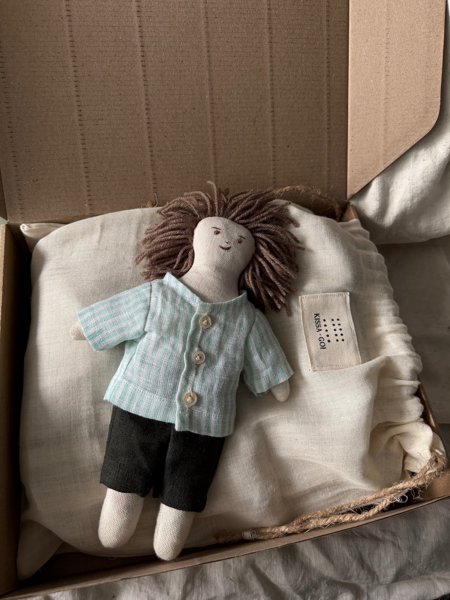 Stuffed doll with a striped shirt and dark pants on a white bag inside a cardboard box. The first friend hand-stitched doll