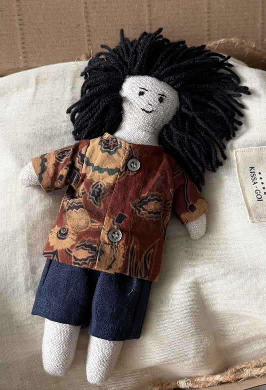 Stuffed doll with black hair, printed linen shirt and dark linen pants in a cardboard box close up.