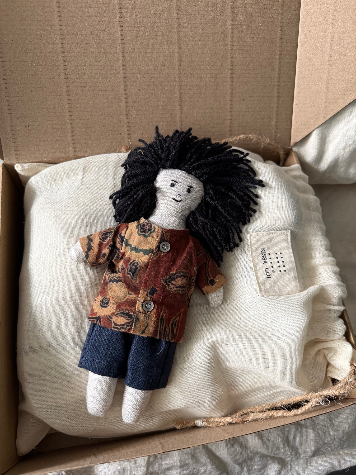 Stuffed doll with black hair, printed linen shirt and dark linen pants in a cardboard box.