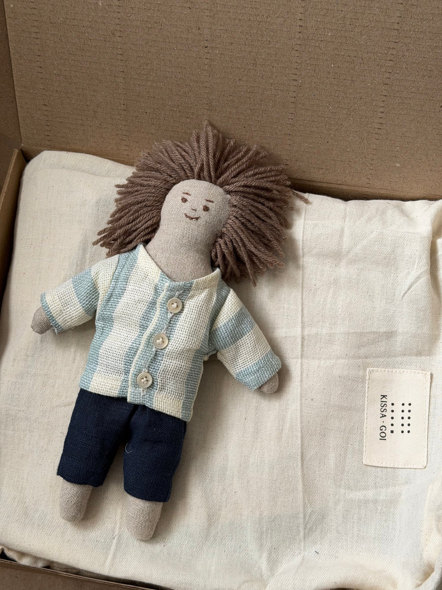 Stuffed doll with a striped shirt and dark pants on a white bag inside a cardboard box. The first friend hand-stitched doll