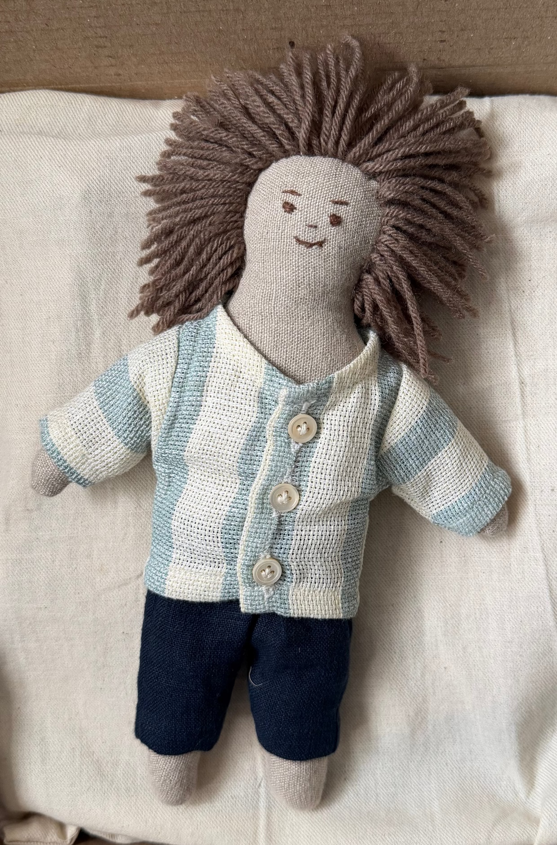 Stuffed doll with a striped shirt and dark pants on a white bag inside a cardboard box. The first friend doll hand-stitched closeup