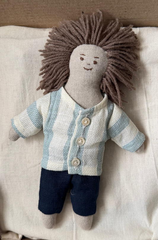 Stuffed doll with a striped shirt and dark pants on a white bag inside a cardboard box. The first friend doll hand-stitched closeup