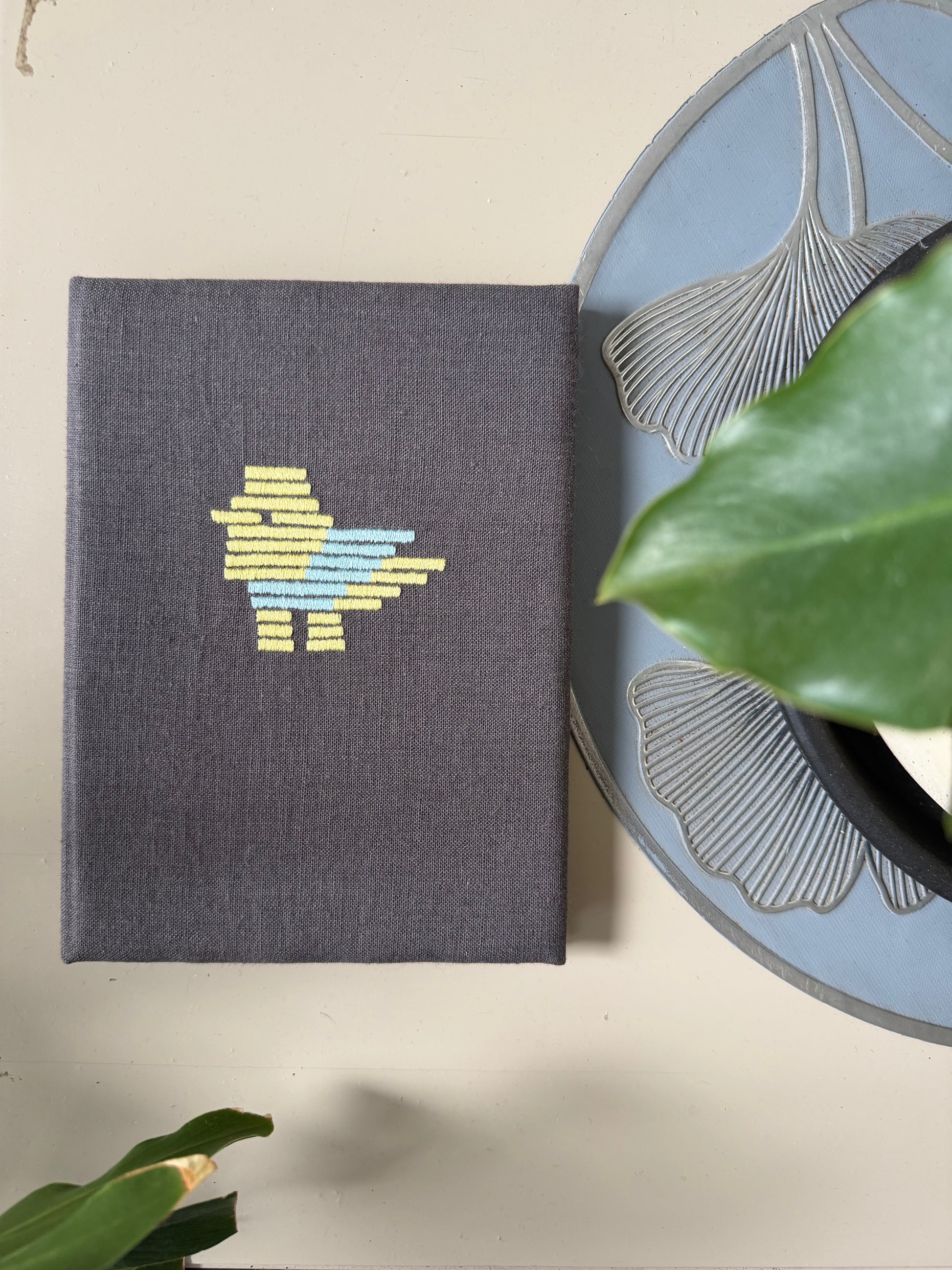 A dark gray linen-covered notebook with a colorful embroidered bird design, styled on a beige surface next to a decorative plate and green leaves.