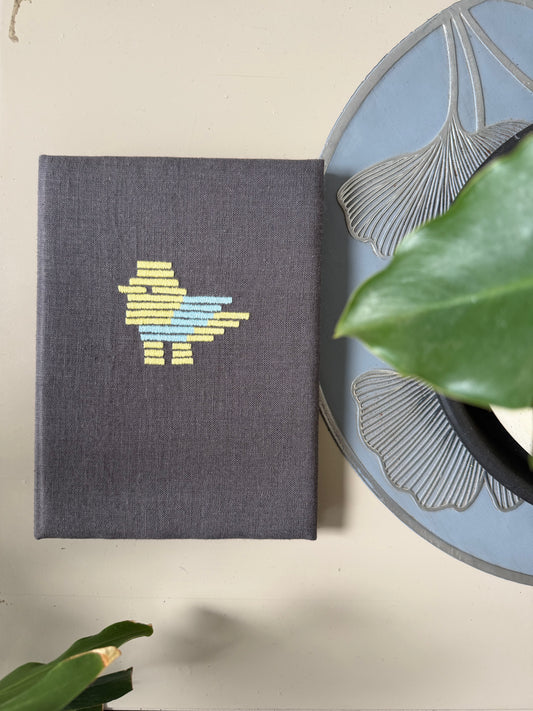 A dark gray linen-covered notebook with a colorful embroidered bird design, styled on a beige surface next to a decorative plate and green leaves.