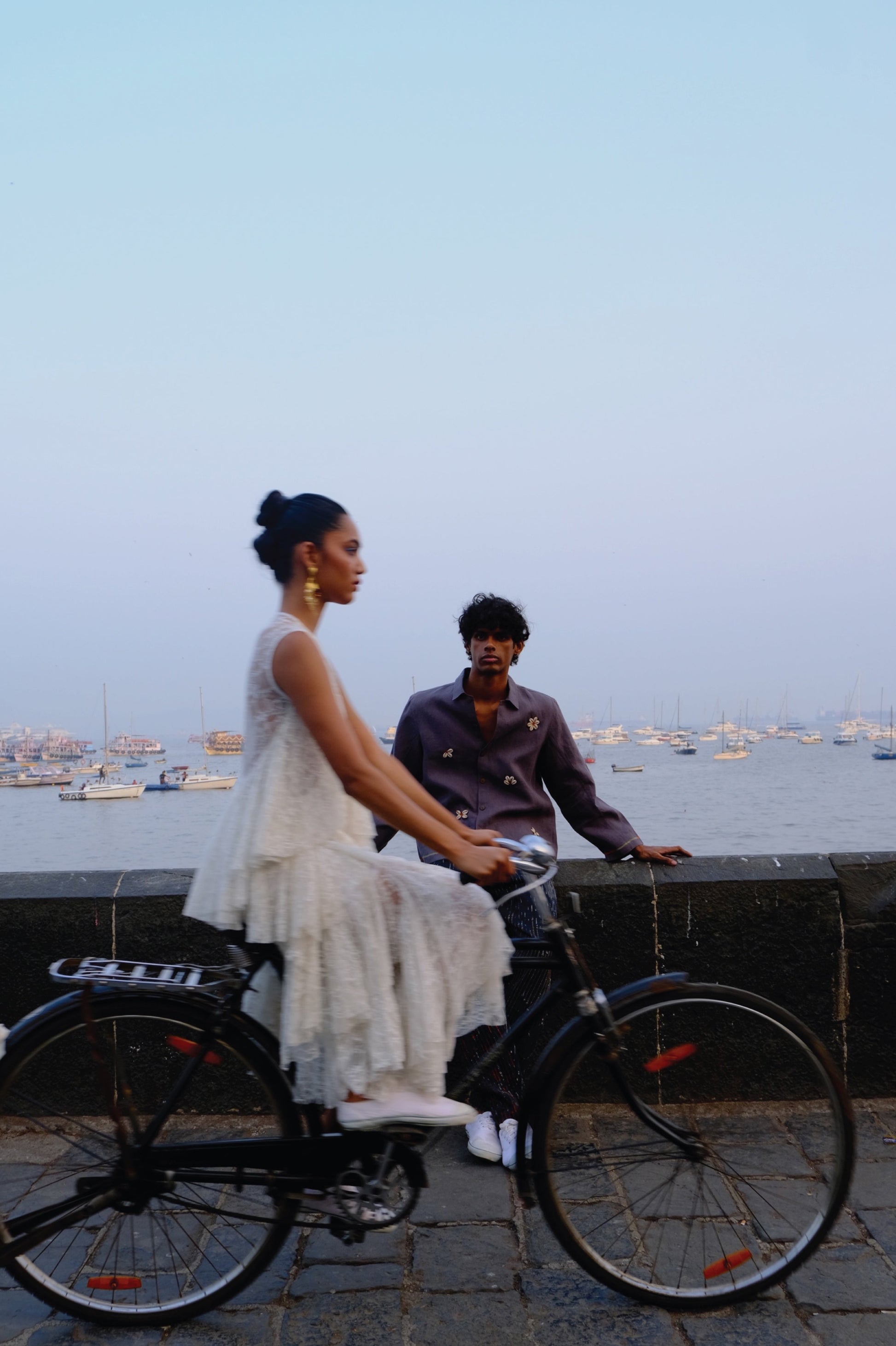 Woman on a bicycle with a man  wearing linen embroidered jacket sitting behind her by a waterfront at dusk.